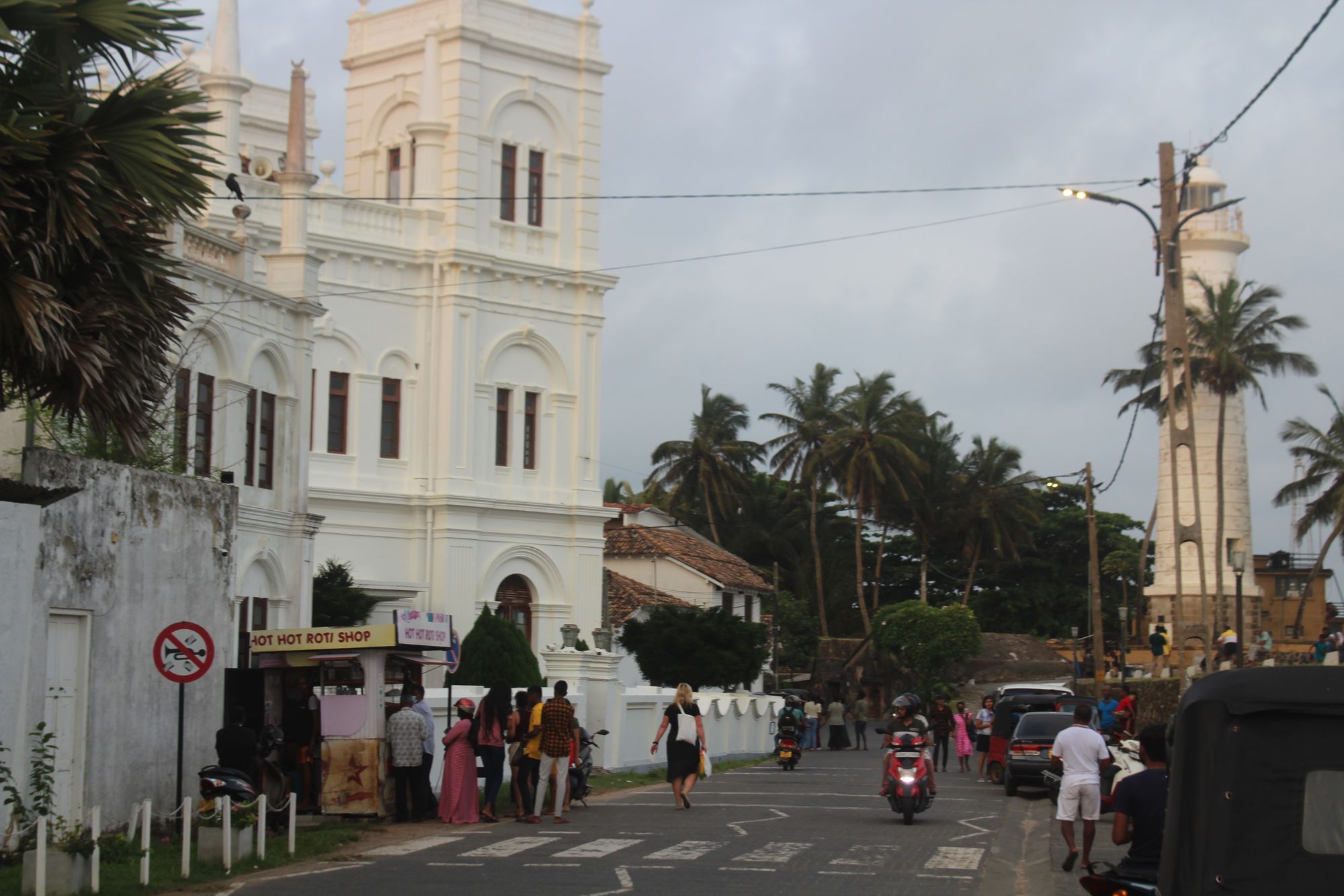 A street in Galle Fort, with people lined up near a street food vendor selling roti, with the Galle Fort lighthouse in the background.